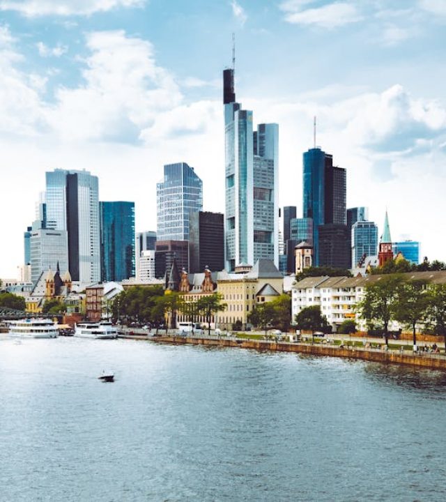 A modern cityscape of Frankfurt with skyscrapers reflecting in the river under a blue sky.